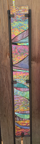 A tall, narrow stained glass panel featuring abstract wavy bands of pink, purple, blue, green, yellow, and orange, framed by dark metal bars against a brown wood background.